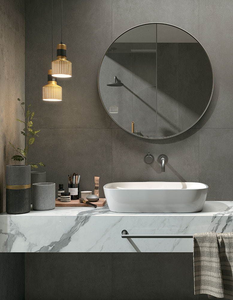 Carrelage en pierre gris 80x80 cm dans une salle de bain aux murs gris, comptoir blanc marbré, accessoires noirs et gris, suspension lumineuse dorée