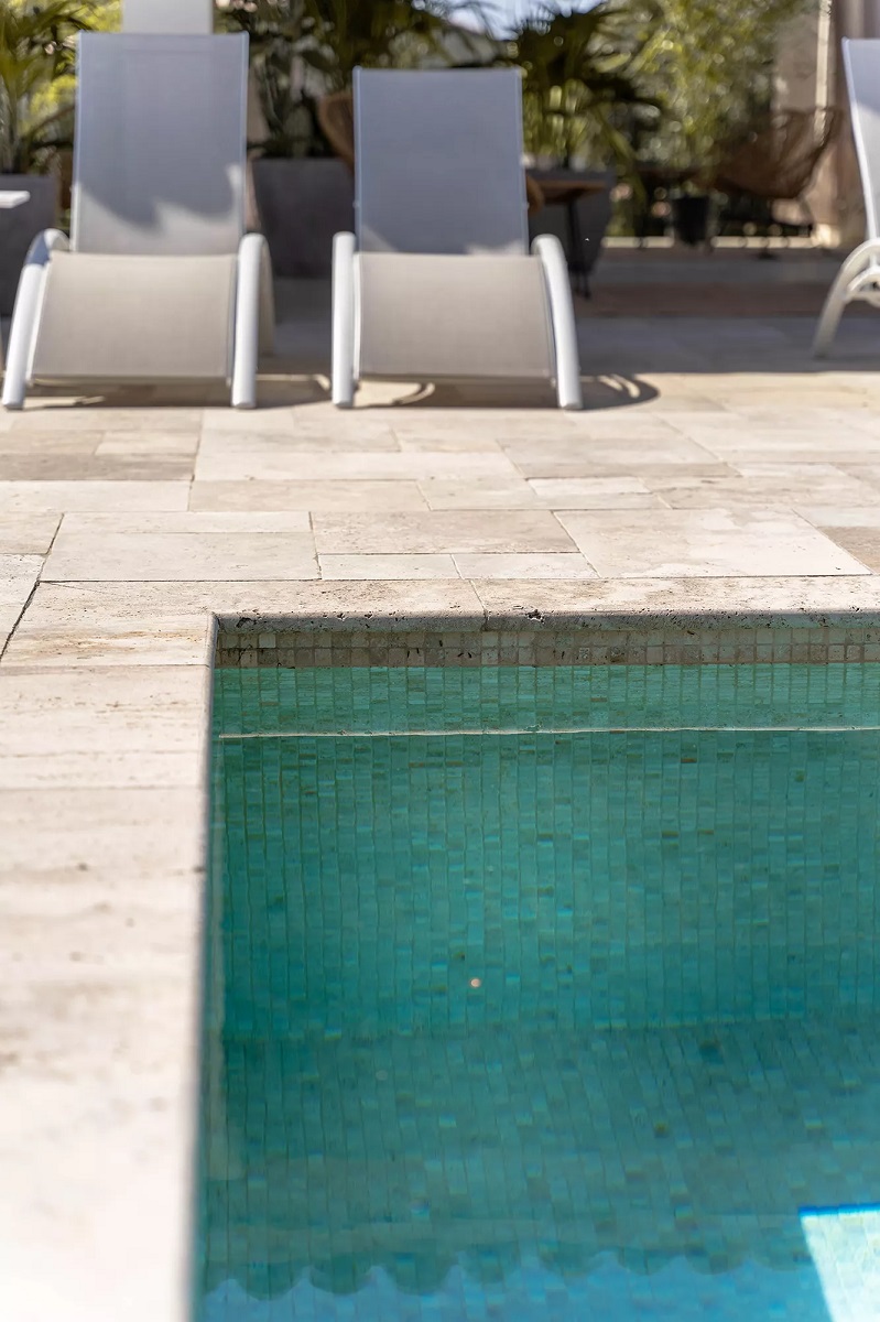Carrelage aspect pierre beige sur bord de piscine extérieure avec chaises longues blanches et gris, végétation en arrière-plan