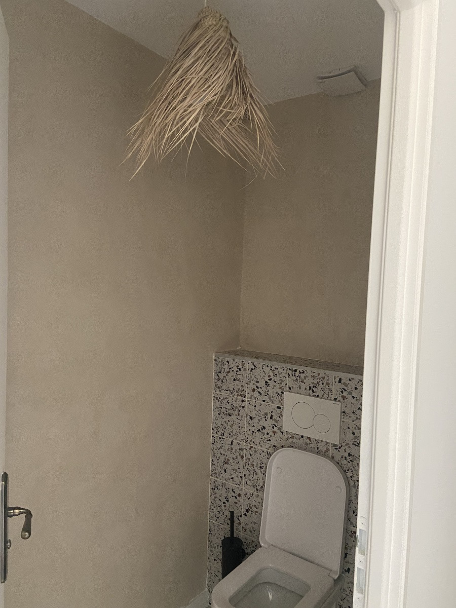 Carrelage Terrazzo blanc avec nuances de gris et noir 20x20 cm dans une salle de bain tons beige avec toilettes et plafonnier en paille