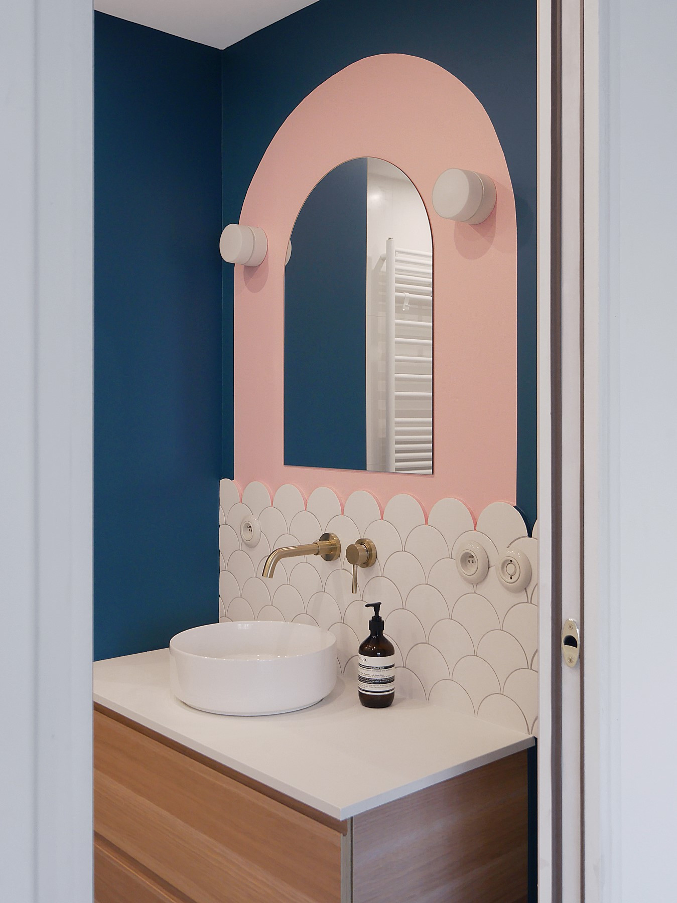 Carrelage uni blanc motif écaille dans une salle de bain aux murs bleu marine et rose, avec vasque blanche et robinetterie dorée