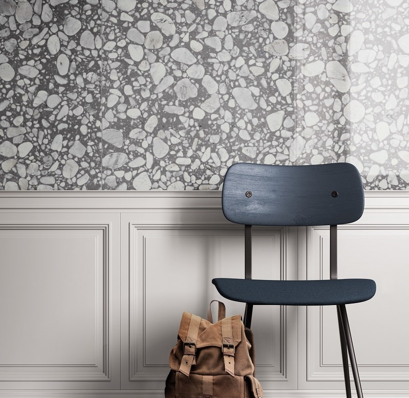 Carrelage Terrazzo gris avec incrustations blanches 60x60 cm sur un mur de salle avec moulures, chaise bleue et sac à dos brun