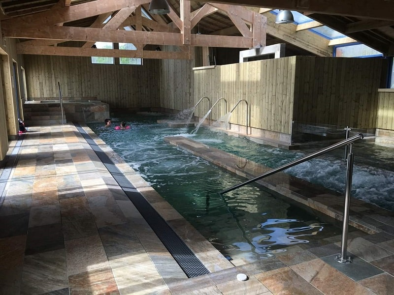 Carrelage aspect pierre beige nuances marron 30x60 cm dans un espace piscine intérieur tons bois clair et métal avec personnes nageant