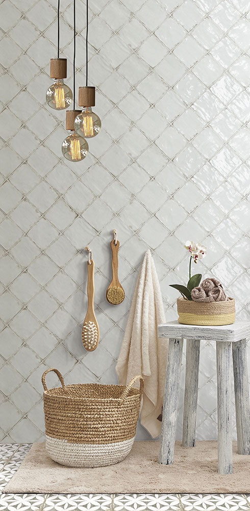 Carrelage Zellige blanc brillant 10x10 cm dans une salle de bain avec panier en osier, serviettes, tabouret en bois et luminaire suspendu