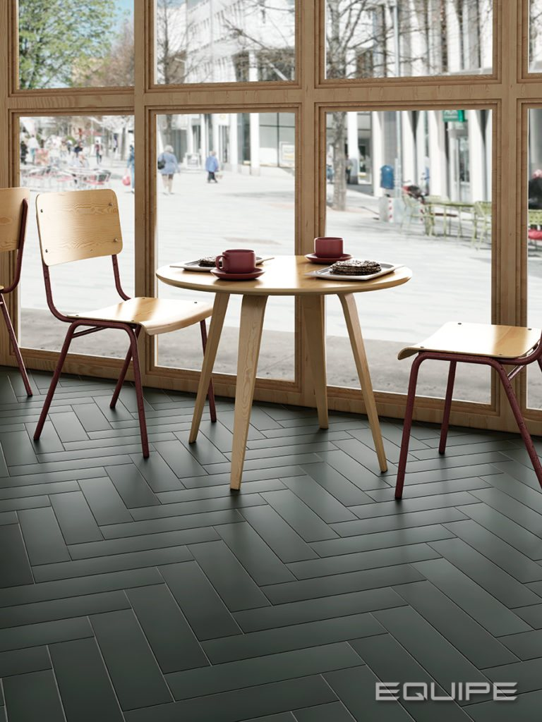 Carrelage uni vert foncé sur le sol dans un café avec tables en bois, chaises rouges et grandes fenêtres donnant sur la rue