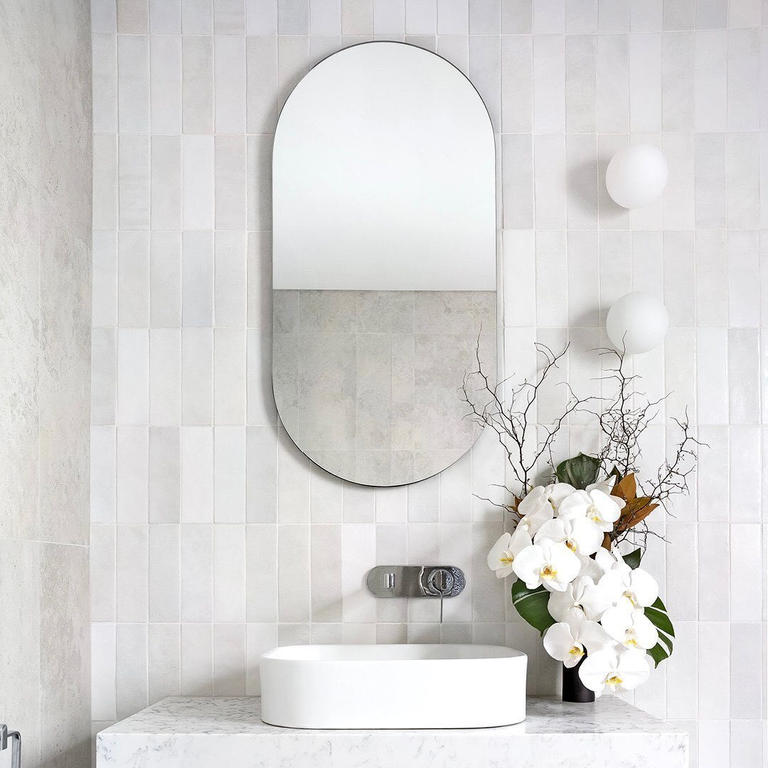 Zellige blanc mat 6,5X20 dans une salle de bain tons de gris et blanc avec vasque, miroir ovale, appliques sphériques et orchidées blanches